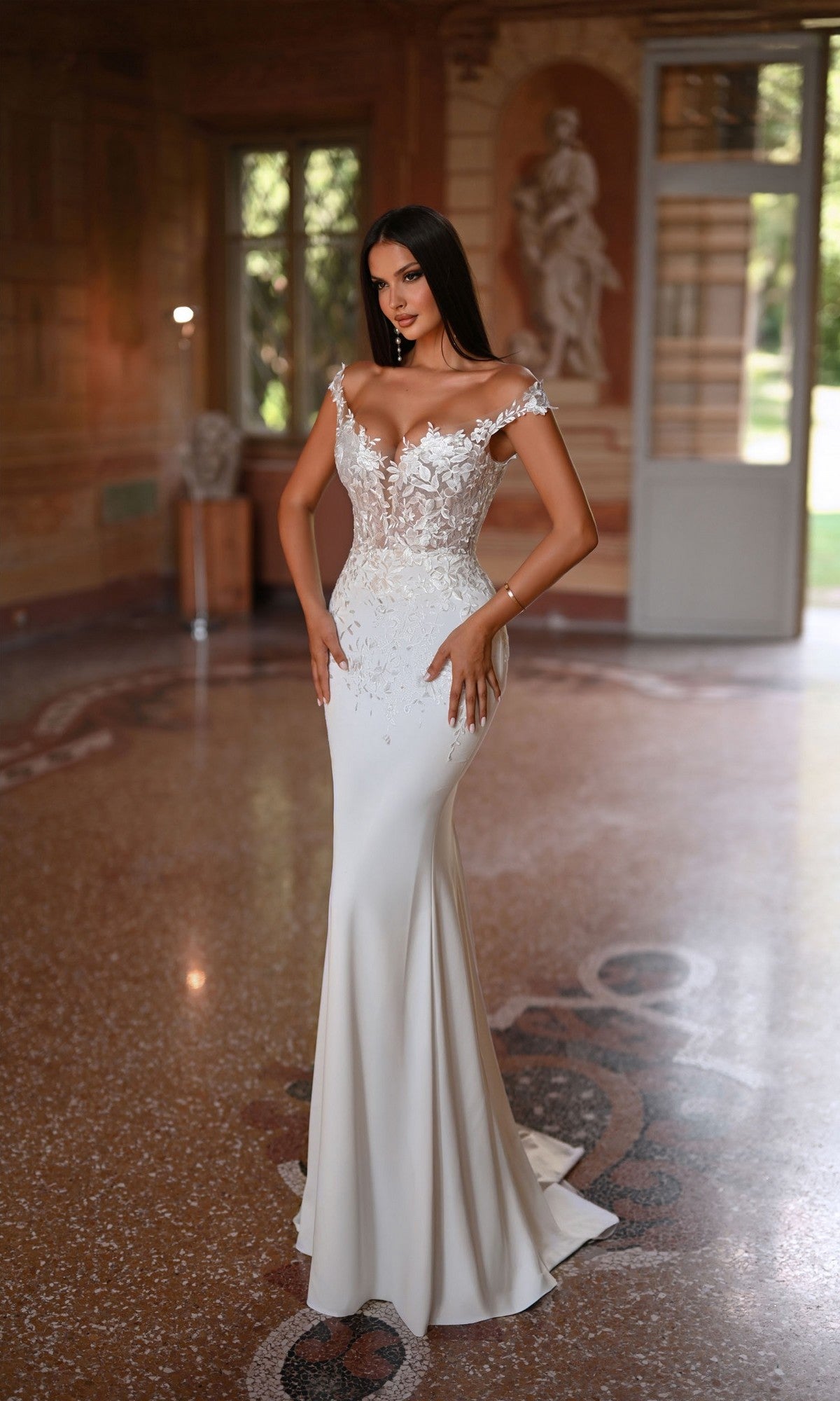 A woman wears the Long White Dress: Alyce 7026—an elegant lace, off-the-shoulder gown—while standing in a grand room with marble floors and classic decor.